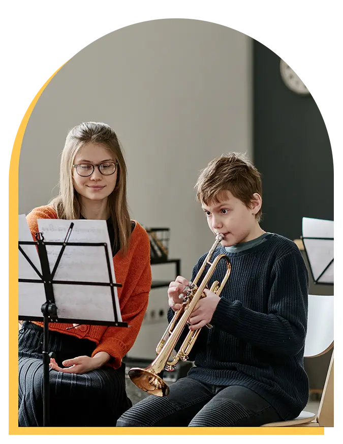 A young boy plays the trumpet while a woman sits beside him, both focused on sheet music during music lessons in a Westchase classroom setting.