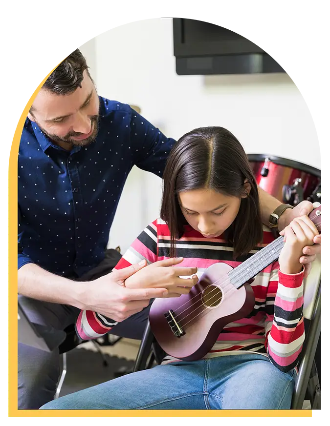 A man teaches a young girl how to play the ukulele during music lessons in Westchase. The girl, wearing a striped shirt, sits and focuses as he guides her hands. A drum set is visible in the background.