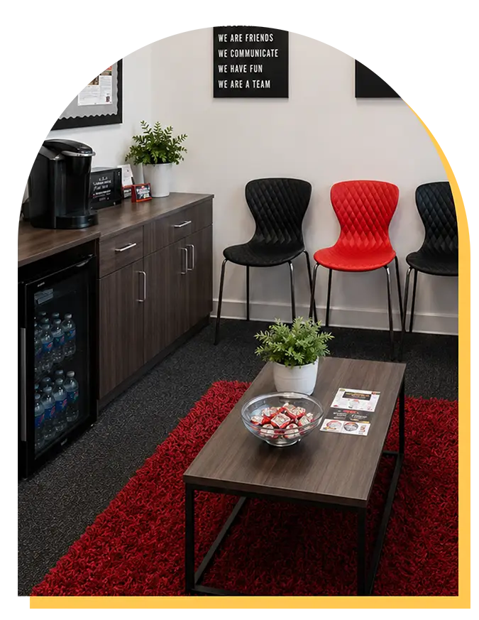 A modern waiting room at Step Up music lessons in Citrus Park features a coffee station, mini fridge, three black chairs, one red chair, snacks and plants on the table, and a red rug. Wall signs inspire teamwork and communication.